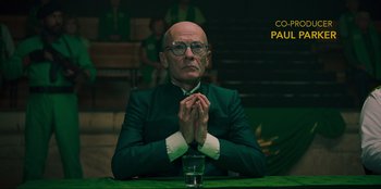 Movie still from “Sniper: G.R.I.T. - Global Response & Intelligence Team” (2023), directed by Oliver Thompson – A man in a green suit sitting in front of a glass; Close Up shot, Over the shoulder angle