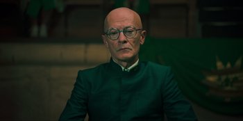 Movie still from “Sniper: G.R.I.T. - Global Response & Intelligence Team” (2023), directed by Oliver Thompson – An older man wearing a green suit and glasses; Close Up shot, Low angle