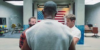 Movie still from “Sniper: G.R.I.T. - Global Response & Intelligence Team” (2023), directed by Oliver Thompson – A group of men standing next to each other in front of an american flag; Medium shot, Over the shoulder angle