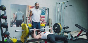 Movie still from “Sniper: G.R.I.T. - Global Response & Intelligence Team” (2023), directed by Oliver Thompson – A man is lifting a barbell in front of another man in a gym; Medium shot, Low angle