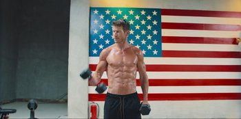 Movie still from “Sniper: G.R.I.T. - Global Response & Intelligence Team” (2023), directed by Oliver Thompson – A man is lifting some weights in front of an american flag; Medium shot, Low angle