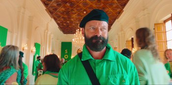 Movie still from “Sniper: G.R.I.T. - Global Response & Intelligence Team” (2023), directed by Oliver Thompson – A man with a beard wearing a green shirt and a black hat; Close Up shot, Low angle