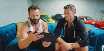 Movie still from “Sniper: G.R.I.T. - Global Response & Intelligence Team” (2023), directed by Oliver Thompson – Two men sitting on a couch looking at a tablet; Medium shot, Over the shoulder angle