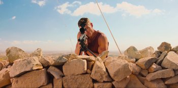 Movie still from “Sniper: G.R.I.T. - Global Response & Intelligence Team” (2023), directed by Oliver Thompson – A man taking a picture with a camera on top of a pile of rocks; Wide shot, Low angle