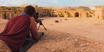 Movie still from “Sniper: G.R.I.T. - Global Response & Intelligence Team” (2023), directed by Oliver Thompson – A man sitting on the ground holding a camera; Wide shot, Over the shoulder angle