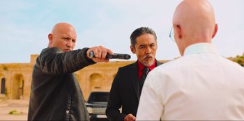 Movie still from “Sniper: G.R.I.T. - Global Response & Intelligence Team” (2023), directed by Oliver Thompson – A man in a black suit and a red tie holding a gun pointed at another man in a black suit and a red tie; Medium shot, Over the shoulder angle