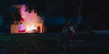 Movie still from “Sniper: G.R.I.T. - Global Response & Intelligence Team” (2023), directed by Oliver Thompson – A woman sitting on the ground in front of a burning house; Extreme Wide shot, Low angle