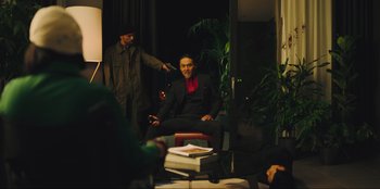 Movie still from “Sniper: G.R.I.T. - Global Response & Intelligence Team” (2023), directed by Oliver Thompson – A man sitting on a chair in front of another man holding a gun; Wide shot, Over the shoulder angle
