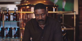 Movie still from “Sniper: G.R.I.T. - Global Response & Intelligence Team” (2023), directed by Oliver Thompson – A man in a brown shirt is sitting in front of a bar; Close Up shot, Low angle