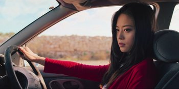 Movie still from “Sniper: G.R.I.T. - Global Response & Intelligence Team” (2023), directed by Oliver Thompson – A woman in a red jacket sitting in the driver's seat of a car; Close Up shot, Low angle
