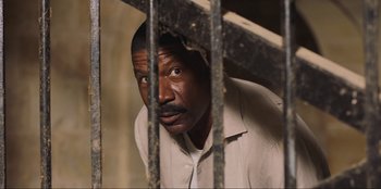 Movie still from “Sniper: G.R.I.T. - Global Response & Intelligence Team” (2023), directed by Oliver Thompson – A man in white shirt behind a metal fence; Close Up shot, High angle
