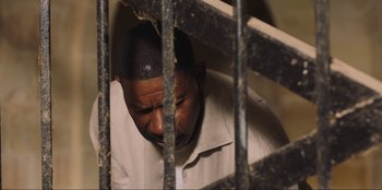Movie still from “Sniper: G.R.I.T. - Global Response & Intelligence Team” (2023), directed by Oliver Thompson – A man in a white shirt is behind bars; Close Up shot, High angle