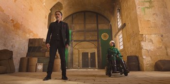 Movie still from “Sniper: G.R.I.T. - Global Response & Intelligence Team” (2023), directed by Oliver Thompson – A man standing next to a man in a wheel chair; Wide shot, Low angle