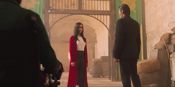 Movie still from “Sniper: G.R.I.T. - Global Response & Intelligence Team” (2023), directed by Oliver Thompson – A woman in a red robe and a man in a black suit stand next to each other in an arched doorway; Wide shot, Over the shoulder angle
