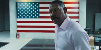 Movie still from “Sniper: G.R.I.T. - Global Response & Intelligence Team” (2023), directed by Oliver Thompson – A man standing in front of an american flag; Medium shot, Low angle