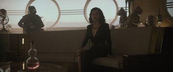 Movie still from “Solo: A Star Wars Story” (2018), directed by Ron Howard – A woman in a black dress sitting on top of a white couch; Medium shot, Over the shoulder angle