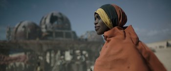 Movie still from “Solo: A Star Wars Story” (2018), directed by Ron Howard – A woman wearing a headscarf is standing in front of a building; Close Up shot, Low angle