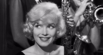 Movie still from “Some Like It Hot” (1959), directed by Billy Wilder – A woman with blonde curly hair and a big smile on her face; Close Up shot, Over the shoulder angle
