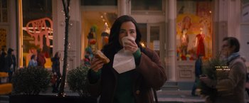 Movie still from “Something from Tiffany's” (2022), directed by Daryl Wein – A woman holding a piece of food while drinking a cup of coffee; Medium shot, Over the shoulder angle