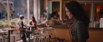 Movie still from “Something from Tiffany's” (2022), directed by Daryl Wein – A woman standing at a counter with a glass dome on top of it; Medium shot, Over the shoulder angle