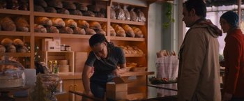 Movie still from “Something from Tiffany's” (2022), directed by Daryl Wein – A woman cutting a loaf of bread in a bakery; Medium shot, Over the shoulder angle