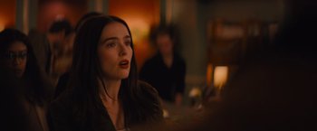 Movie still from “Something from Tiffany's” (2022), directed by Daryl Wein – A woman sitting in front of a table with other people in the background; Close Up shot, Over the shoulder angle