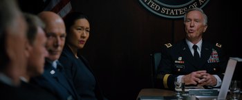 Movie still from “Sonic the Hedgehog” (2020), directed by Jeff Fowler – A woman sitting at a table in front of an american flag; Medium shot, Over the shoulder angle