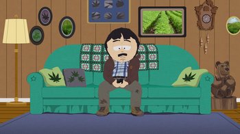 Movie still from “South Park: Joining the Panderverse” (2023), directed by Trey Parker – A man sitting on a green couch holding a remote control; Wide shot, High angle