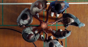 Movie still from “Space Jam” (1996), directed by Joe Pytka – A group of men standing on a basketball court holding a ball; Wide shot, Overhead angle