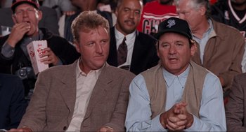 Movie still from “Space Jam” (1996), directed by Joe Pytka – A couple of men sitting next to each other in a crowd; Medium shot, Over the shoulder angle