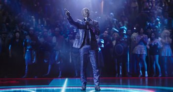 Movie still from “Space Jam: A New Legacy” (2021), directed by Malcolm D. Lee – A man in a blue suit is holding a microphone; Wide shot, Over the shoulder angle