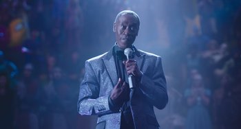 Movie still from “Space Jam: A New Legacy” (2021), directed by Malcolm D. Lee – A man holding a microphone and wearing a suit and tie; Medium shot, Low angle