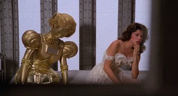 Movie still from “Spaceballs” (1987), directed by Mel Brooks – A woman in a white dress sitting next to a gold statue; Medium shot, Low angle