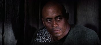 Movie still from “Spartacus” (1960), directed by Stanley Kubrick – A bald man with a beard and a chain around his neck; Close Up shot, Low angle