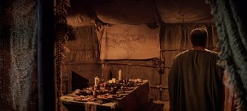 Movie still from “Spartacus” (1960), directed by Stanley Kubrick – A table set up for a meal in a hut style building; Medium shot, Over the shoulder angle