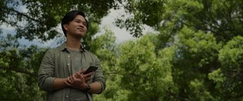 Movie still from “Spiderhead” (2022), directed by Joseph Kosinski – A man holding a cell phone while standing next to a tree; Medium shot, Low angle