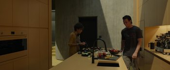 Movie still from “Spiderhead” (2022), directed by Joseph Kosinski – A woman standing at a counter in front of some food; Wide shot, High angle