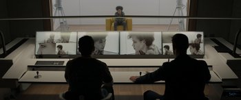 Movie still from “Spiderhead” (2022), directed by Joseph Kosinski – A group of people sitting in front of a large screen tv; Wide shot, Over the shoulder angle
