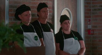 Movie still from “Spin Me Round” (2022), directed by Jeff Baena – A group of people wearing aprons standing in front of a mirror; Medium shot, Over the shoulder angle