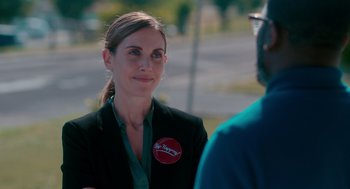 Movie still from “Spin Me Round” (2022), directed by Jeff Baena – A woman in a black jacket and a red button; Close Up shot, Over the shoulder angle