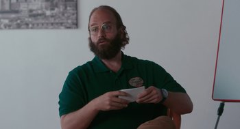 Movie still from “Spin Me Round” (2022), directed by Jeff Baena – A man sitting in a chair holding a nintendo wii game controller; Medium shot, Low angle