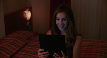 Movie still from “Spin Me Round” (2022), directed by Jeff Baena – A beautiful woman sitting on a bed looking at a tablet computer; Close Up shot, Over the shoulder angle