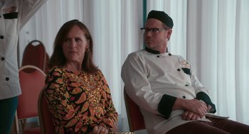 Movie still from “Spin Me Round” (2022), directed by Jeff Baena – A man and a woman sitting next to each other; Medium shot, Over the shoulder angle