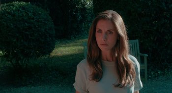 Movie still from “Spin Me Round” (2022), directed by Jeff Baena – A beautiful woman with long brown hair sitting on a bench; Close Up shot, Over the shoulder angle