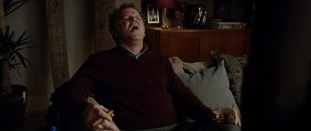 Movie still from “Carnage” (2011), directed by Roman Polanski – A man sitting on top of a couch while holding a cigarette; Medium shot, High angle