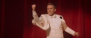 Movie still from “Spirited” (2022), directed by Sean Anders – A man in a white suit and bow tie holding his fist in the air; Medium shot, High angle