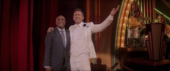 Movie still from “Spirited” (2022), directed by Sean Anders – A man in a white suit standing next to another man in a gray suit; Medium shot, Over the shoulder angle