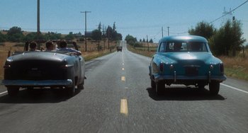 Movie still from “Stand by Me” (1986), directed by Rob Reiner – Two vintage cars driving down the road in the middle of the day; Wide shot, Low angle