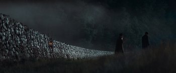A man in a black coat and a black hat walking on a grassy hill