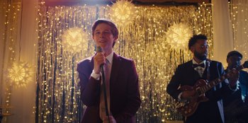 Movie still from “Stargirl” (2020), directed by Julia Hart – A man in a suit and tie holding a microphone in front of lights; Medium shot, Over the shoulder angle